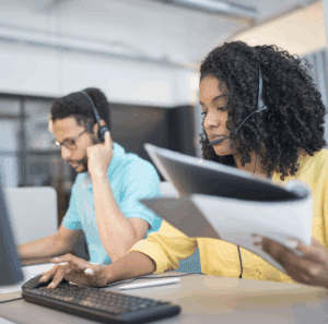 A woman examines a tablet while her colleague uses a headset and a desktop computer in an office setting.