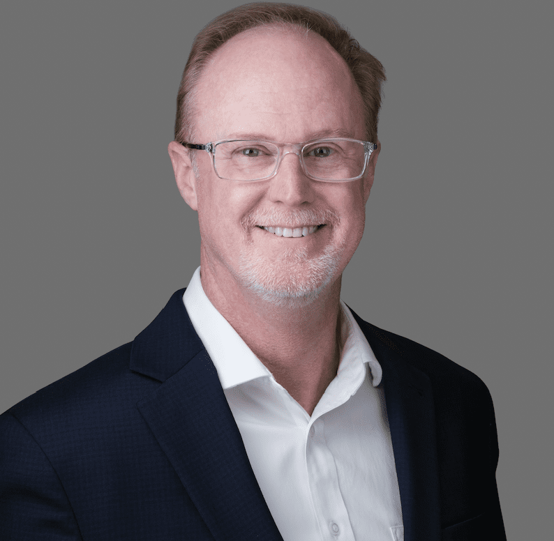 A smiling individual wearing glasses and dressed in a professional blue jacket over a white shirt poses against a neutral gray background.