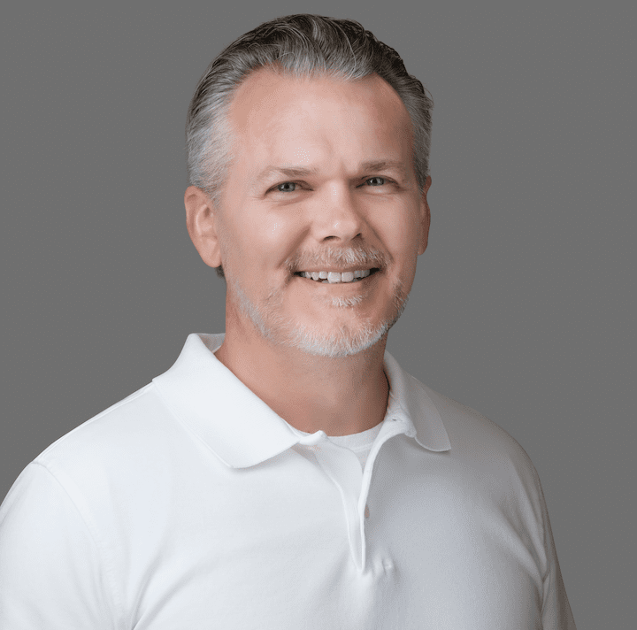 A smiling individual with graying hair, wearing a white polo shirt, presents a friendly and approachable demeanor against a gray background.