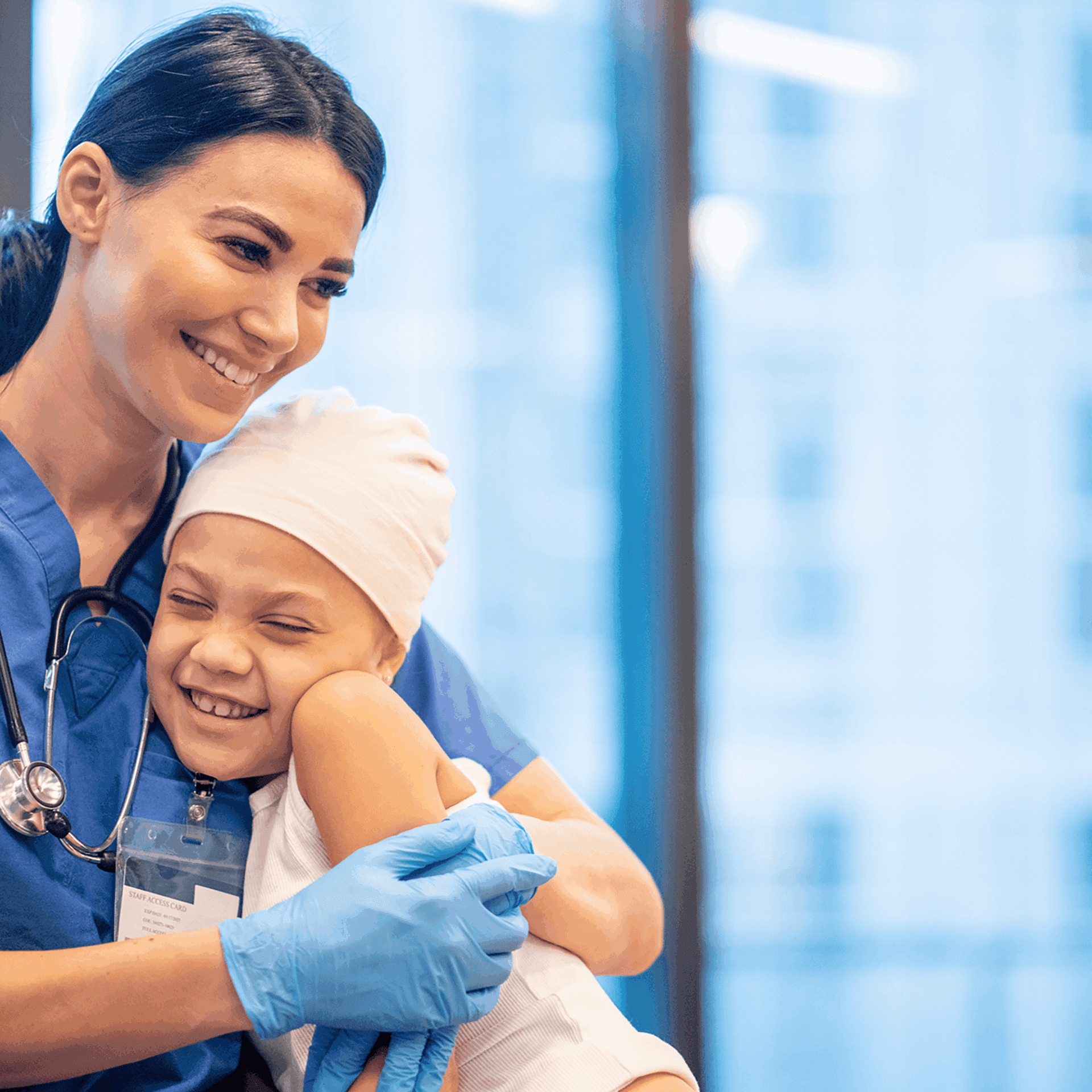 A healthcare worker in blue scrubs warmly embraces a young patient wearing a headscarf in a hospital setting.