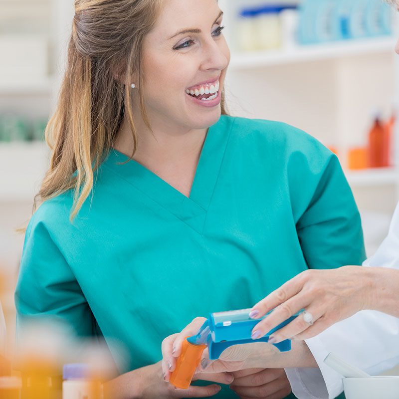 A smiling healthcare professional in teal scrubs is actively engaged in reading a document in a brightly lit pharmacy.