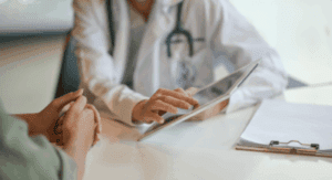 A doctor in a white coat with a stethoscope is using a tablet during a consultation.