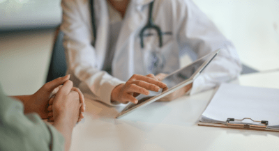 A doctor in a white coat with a stethoscope is using a tablet during a consultation.