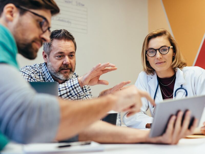 A group of professionals is engaged in a discussion around a table, with one of them gesturing and another looking at a digital tablet.