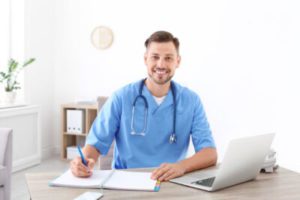 A cheerful healthcare professional in scrubs sits at a desk, writing notes and using a laptop.