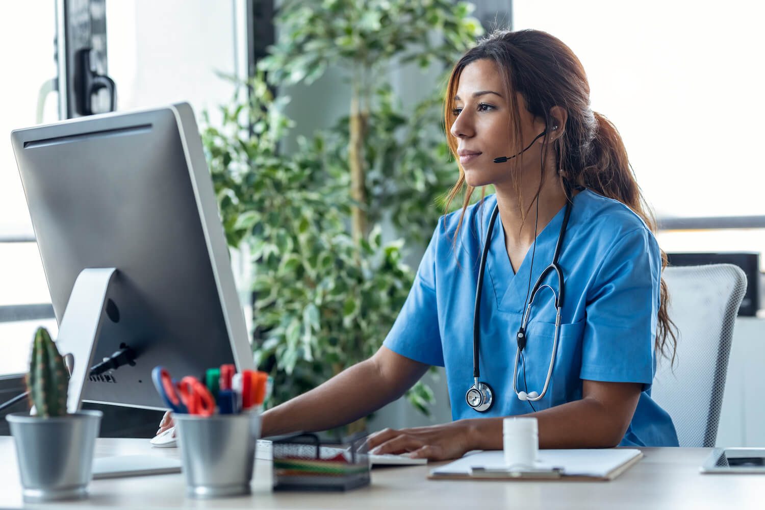 A healthcare professional in blue scrubs and a headset sits at a desk, working intently on a computer in a bright office setting.