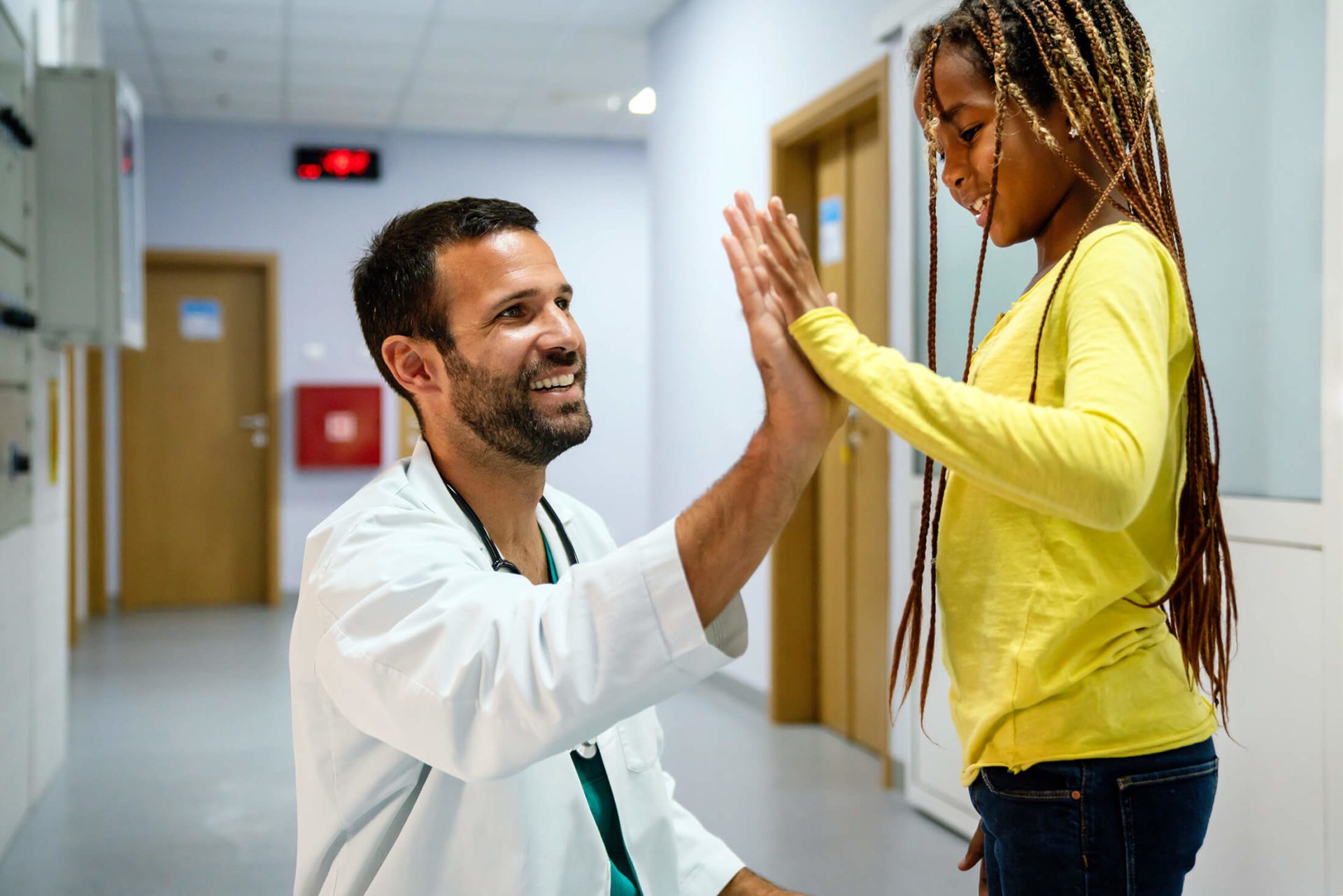 A smiling doctor in a white coat gives a high-five to a young patient in a yellow shirt inside a hospital hallway.