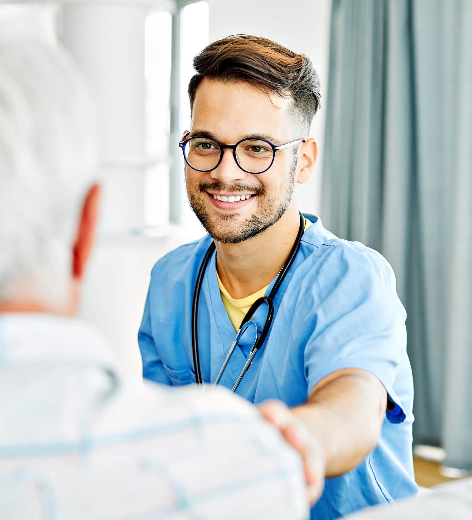 A smiling healthcare professional wearing glasses and a blue scrub shakes hands with a patient in a clinical setting.