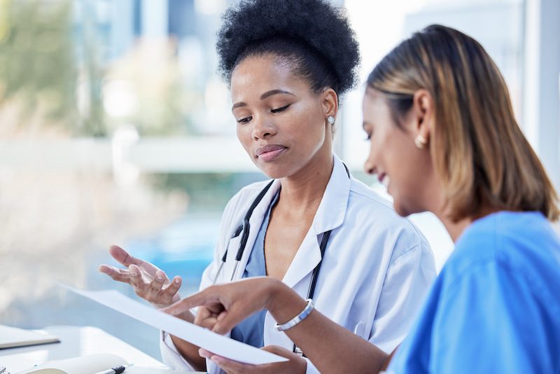 A doctor in a white coat is consulting a clipboard with a colleague in a brightly lit hospital setting.